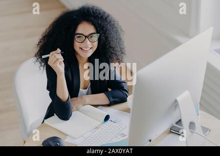 Une femme afro-américaine heureuse à peau foncée tient le stylo, écrit quelques informations, travaille comme éditeur, regarde avec joie l'appareil photo, tient le stylo, utilise la moder Banque D'Images
