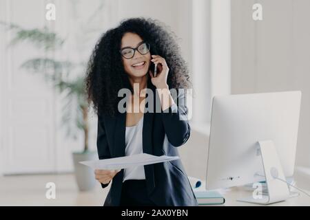 Le Manager féminin chargé tient un téléphone portable près de l'oreille, tient des documents, vérifie le rapport, a une conversation téléphonique, porte des lunettes et une tenue élégante, sourit largement Banque D'Images