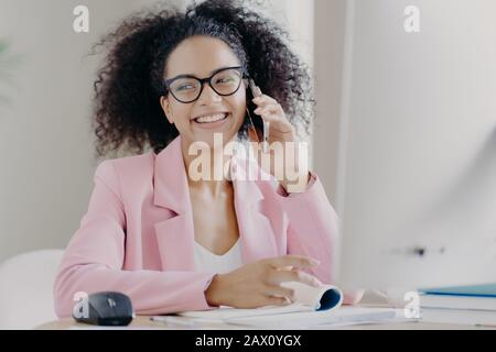 Une femme épluchée sombre à poil bouclés aime discuter par téléphone avec un collègue, a une expression heureuse, se trouve sur le bureau, porte des lunettes et un sui rose Banque D'Images