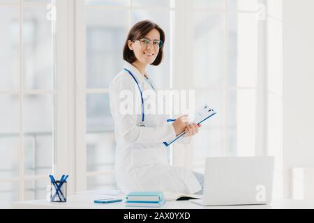 Femme médecin assis sur la table, écrit des informations dans le presse-papiers, pose sur le lieu de travail dans le bureau de l'hôpital, travaille avec ordinateur portable, sourires agréablement Banque D'Images