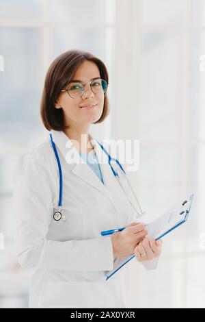 Un expert en cardiologie qualifié avec stéthoscope prépare un séminaire sur la santé, donne des conseils sur les premiers soins, écrit des informations dans le presse-papiers, porte Banque D'Images