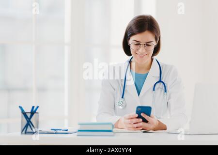 Le travailleur médical féminin utilise un smartphone, lit des conseils pour guérir la maladie sur Internet, pose dans un bureau médical, porte un uniforme médical spécial, voit le resu Banque D'Images