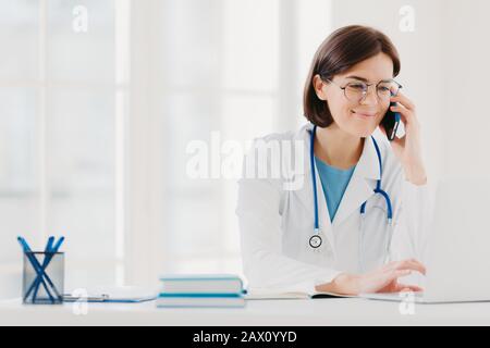 Thérapeute souriant focalisé sur l'écran de l'ordinateur portable, les appels via un smartphone moderne, porte une robe de médecin, lunettes transparentes, se trouve dans l'espace de travail, Banque D'Images