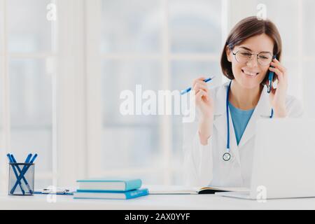 Photo de la femme heureuse thérapeute ou médecin a une conversation téléphonique avec le patient, donne des conseils pour guérir la maladie, porte la robe blanche, lit l'information Banque D'Images