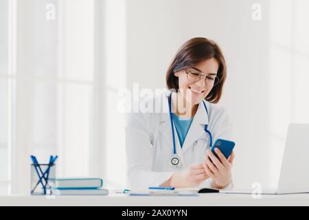 Une femme médicale positive impliquée dans le chat, tient un téléphone mobile et envoie des messages, porte une robe médicale, a une expression chaleureuse et gaie, pose Banque D'Images