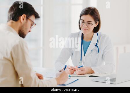 Le patient masculin écrit le formulaire de demande, obtient des conseils et des conseils sur le traitement à domicile du médecin, utiliser l'ordinateur portable et le document médical au bureau, pose Banque D'Images