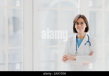 Un médecin professionnel confiant se tient debout avec des bras croisés, porte une robe médicale blanche avec stéthoscope, pense au travail positivement, pose contre bi Banque D'Images