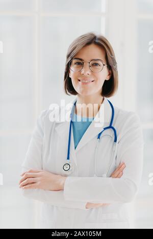 Beau médecin gai femme porte robe blanche, lunettes et phonendoscope, garde les mains croisées, regarde en toute confiance l'appareil photo, a des cheveux courts et sombres, St Banque D'Images