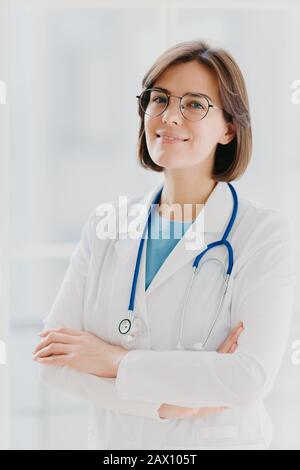 Tir de brunette confiante femme médecin ou chirurgien se tient avec les mains croisées, porte l'uniforme blanc, se soucie de la santé des patients prêts à donner advic Banque D'Images