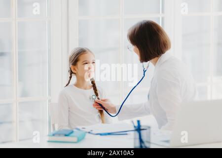 Femme pédiatre donne la consultation à la petite fille, parler de la santé et des symptômes, écoute le coeur avec phonendoscope, fait la prescription, pose dans le dos Banque D'Images