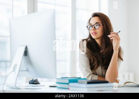 Une dame aux cheveux sombres, centrée sur le grand moniteur, assise sur le bureau, tient le stylo et écrit des notes, porte des lunettes pour la correction de la vision, pose dans le spa de collègue Banque D'Images