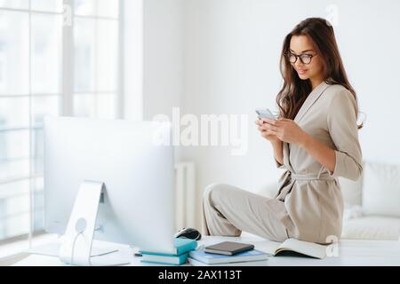Une élégante femme en costume beige lit les nouvelles sur le site Web attentivement, se trouve sur le bureau seul dans le cabinet, le moniteur d'ordinateur et les blocs-notes autour. Femme e Banque D'Images