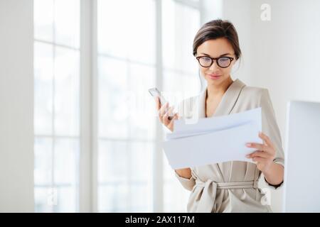 L'employé féminin satisfait, concentré dans les documents, lit attentivement le rapport préparé, tient un smartphone moderne, porte des lunettes transparentes et un sui d'affaires Banque D'Images