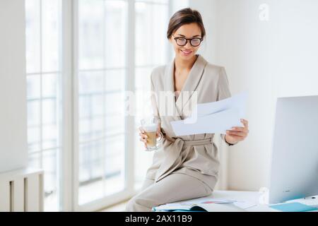 Une femme heureuse aux cheveux sombres, souriante, vérifie les factures de documents, déguisée costume beige, boit des boissons, se trouve sur le bureau près du grand moniteur, tient Banque D'Images