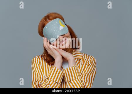 Jolie femme à tête rouge porte les yeux bandés, maintient les mains enfoncées près du visage, voit des rêves agréables tout en dormant, vêtu de vêtements domestiques Banque D'Images