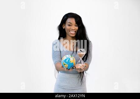 Jeune fille africaine souriante en robe grise tient dans les mains monde de la Terre globe et sablier, isolé sur fond blanc. Entretien de la planète, sauver la terre, non Banque D'Images