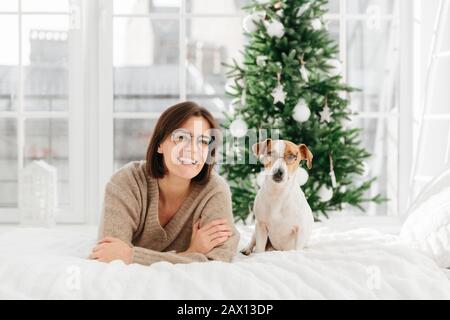 Une femme joyeuse avec des cheveux sombres courts porte des lunettes et des verres ronds, un chien drôle dans des lunettes pose près de l'hôte, se trouve sur le lit, ont un bon repos pendant Chris Banque D'Images