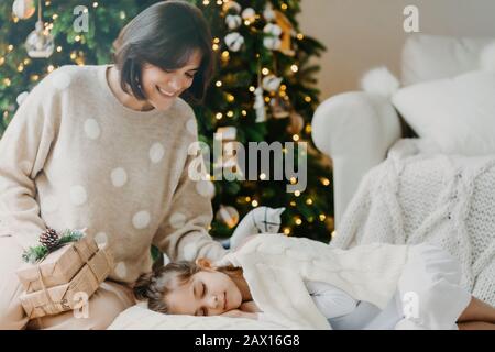 Profitez de Noël à la maison. Affectueuse jeune mère pose près de la fille dormant, va faire la surprise, tient des cadeaux présents, croire en des miracles. Magie Banque D'Images