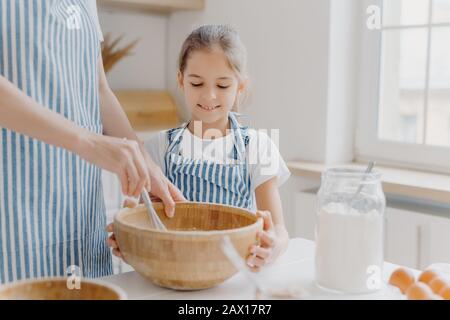 Curieuse petite aide féminine regarde attentivement comment la mère cuit, aide à battre les ingrédients, porte un t-shirt blanc et un tablier rayé, prépare un gâteau savoureux Banque D'Images