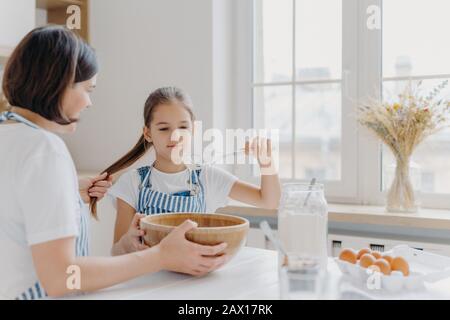 Une petite aide pour enfants fouille la crème avec le batteur, apprend à cuire, écoute des conseils utiles de la mère qui tient sa queue de poney, s'asseoir dans la cuisine blanche Banque D'Images