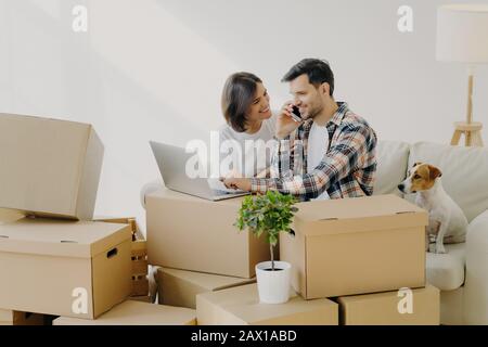 Heureux mari et femme se déplacent dans la nouvelle maison achetée, acheter des meubles en ligne, utiliser la technologie moderne, l'homme surfs Internet par ordinateur portable et parler par téléphone, assis Banque D'Images