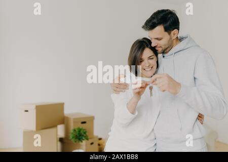 Enchanté femme et homme propriétaires de maison aiment acheter nouveau plat, les clés de tenir fromm leur propre appartement, poser contre le mur blanc avec des boîtes de cardboad non emballées. Banque D'Images