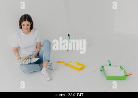 La femme satisfaite brunette choisit la couleur des échantillons, allant à la salle de remise à neuf, se trouve au sol dans des vêtements décontractés, entourés de plateaux et de rouleaux de peinture Banque D'Images