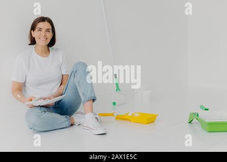 Photo horizontale de la femme souriante choisit la meilleure couleur pour l'appartement, tient la palette, vêtu de vêtements décontractés, peint les murs, rénove sa maison Banque D'Images