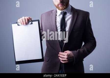 Studio portrait d'un jeune homme barbu de vingt-cinq, dans un costume officiel, tenant un dossier avec une feuille de lecture pour le texte. Espace libre Sur un g Banque D'Images