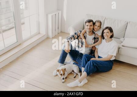 Belle famille européenne de père, de mère et de leur fille assis au sol près d'un canapé dans une grande chambre blanche, le chien pedigree se trouve à proximité, ont jour de déménagement, ch Banque D'Images