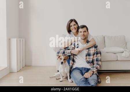 Le portait d'une femme affectueuse embrasse le mari qui se trouve au sol près du canapé, leurs poses d'animal de compagnie préférées à l'appareil photo, faire de la photo, être à la maison, acheter un nouveau point d'accès Banque D'Images