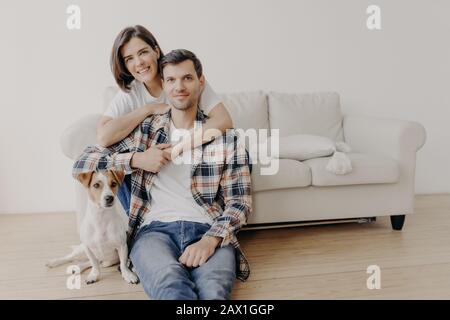 Photo d'une femme heureuse embrasse son mari, pose sur un canapé, petit chien assis au sol près du propriétaire, regarder tout à l'appareil photo avec des expressions heureuses, ont romantis Banque D'Images