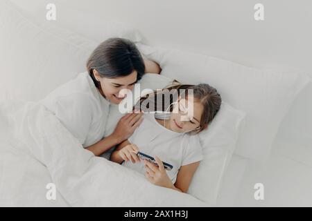 La photo d'une mère heureuse passe du temps libre avec une petite fille charmante qui se concentre sur le smartphone, écoute des chansons dans les écouteurs, pose dans le lit avec un doux blanc Banque D'Images