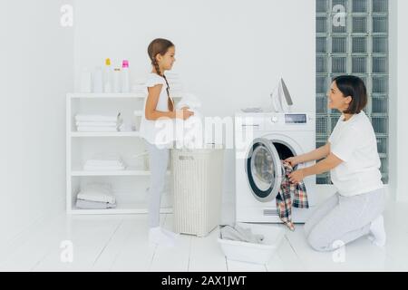 Content mère et fille occupés à faire la lessive à la maison, ont des visages heureux, femme de brunette charge machine à laver, petite fille se tient près du panier avec blanc Banque D'Images