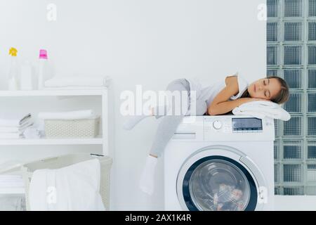 La photo horizontale de la petite fille a le repos sur la machine à laver, se sent fatigué après avoir aidé maman au sujet de la maison, dort sur la machine à laver pose dans la buanderie avec la clim Banque D'Images