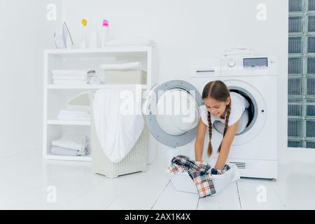 Jolie petite fille femme de ménage pose à l'intérieur de la machine à laver, prend chemise à carreaux de bassin, engagé dans la lessive, a l'expression heureuse, deux lits Banque D'Images