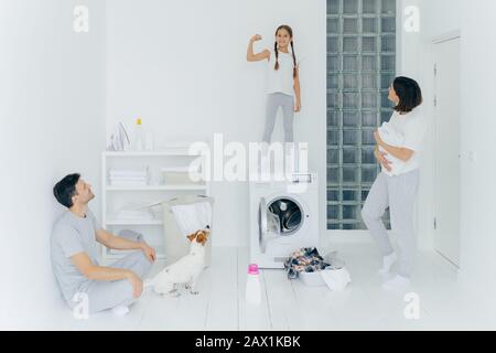 Une jeune fille forte et gaie lève le bras, montre des biceps, amuse-guette les parents, se brisent après le lavage des vêtements, pose dans une grande buanderie blanche avec animal de compagnie, lavage Banque D'Images