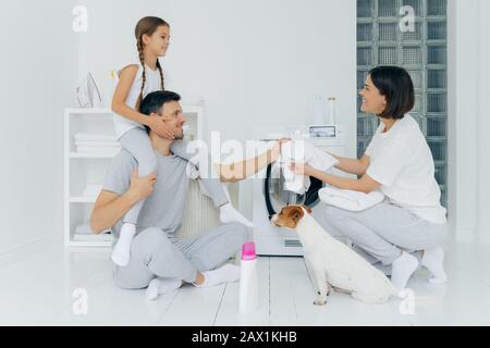 Famille, ménage, concept de lavage. Le père souriant donne une balade en porgyback à la petite fille donne à la femme un t-shirt blanc à laver assis sur le sol au lavage Banque D'Images