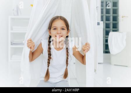 Heureuse fille européenne avec deux plis, poses près de vêtements cheval entre le linge blanc de séchage, pose dans la salle de lavage contre fond flou, le blanc colo Banque D'Images