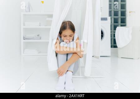 Une petite fille triste et attentionnée se trouve au sol dans la salle à laver près du sèche-linge, porte un pantalon et des chaussettes blanches, un lave-linge, un panier à linge et Banque D'Images