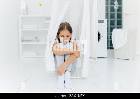 Petite fille adorable assise sur le sol, punie par les parents, pose près du sèche-linge, concentré avec une triste expression, machine à laver, panier avec Banque D'Images