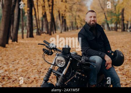 Le motocycliste barbu fait du vélo noir, tient le casque, voyage sur son propre transport, pose en parc pendant la saison d'automne, regarde avec plaisir la caméra. Banque D'Images