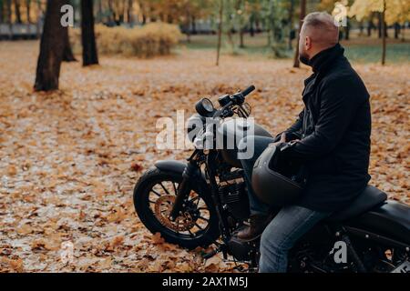 Vue arrière du biker masculin barbu semble soigneusement quelque part dans la distance, pose sur moto noire, tient casque de protection, passe du temps libre dans Banque D'Images