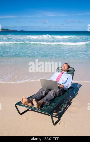 Homme d'affaires pieds nus travaillant sur son ordinateur portable sur une chaise de plage se reposant sur la rive d'une plage tropicale vide Banque D'Images