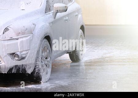 Modelage avec mousse sur la voiture dans le lavage de voiture en libre-service. Voiture blanche à la station de lavage de voiture. De l'eau et du savon blanc coule. Banque D'Images