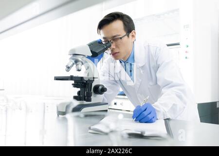 Scientifique asiatique portant un pelage blanc examinant l'échantillon au microscope, portrait horizontal Banque D'Images