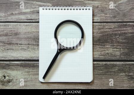 Le Bloc-notes avec une feuille blanche vierge dans un papier de vérification se trouve avec une loupe sur l'arrière-plan des planches en bois. Banque D'Images