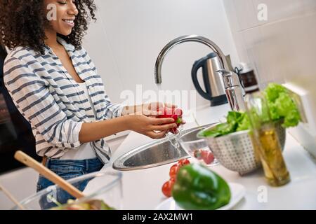 Dinenr. Sain Jeune femme africaine debout dans la cuisine à la maison laver les légumes souriant gai gros plan Banque D'Images