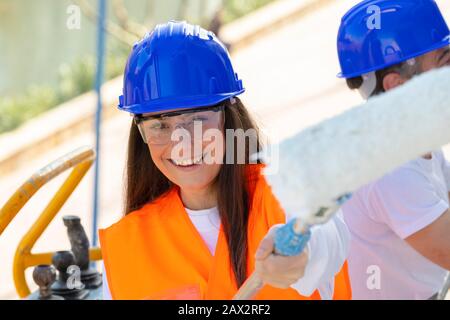 Gros plan d'une belle femme portant un casque de sécurité et des lunettes et tenant un rouleau de peinture souriant à l'appareil photo sur un arrière-plan hors de la mise au point. Travail c Banque D'Images