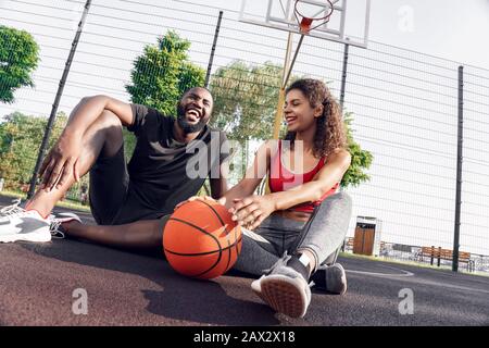 Activité En Plein Air. Couple africain assis avec ballon sur le terrain de basket-ball en riant la vue de fond heureuse Banque D'Images